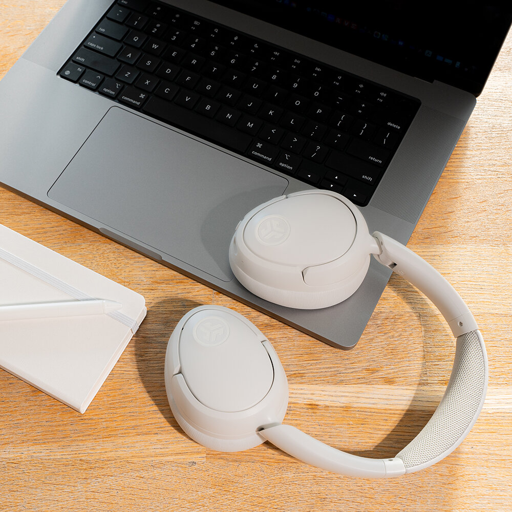 Białe słuchawki na laptopie. Z lewej strony notatnik i długopis. Tłem jest drewniana powierzchnia.