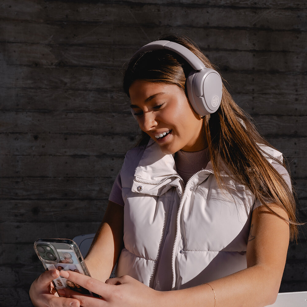 Kobieta w słuchawkach i kamizelce uśmiecha się, patrząc na telefon. Betonowa ściana w tle.