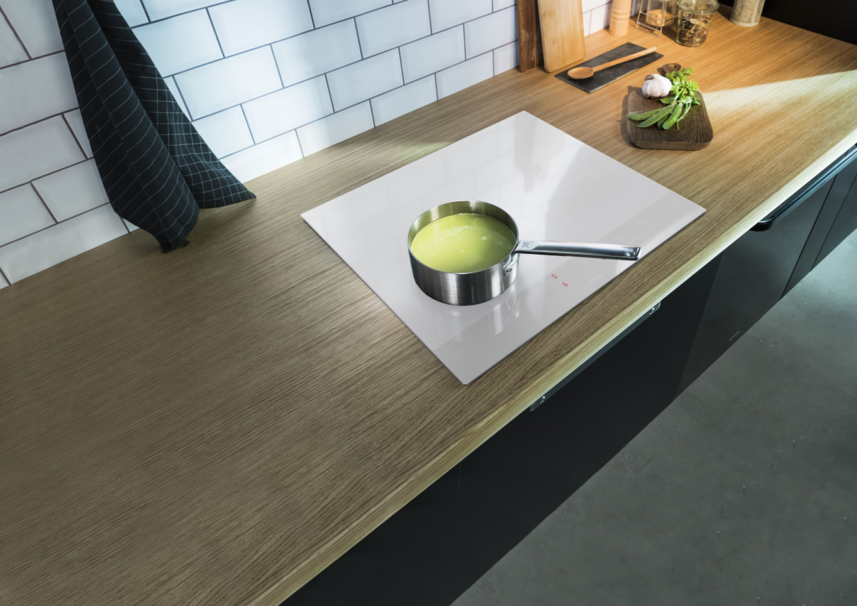 Scena kuchenna z garnkiem zupy na białej płycie grzewczej. Drewniany blat z czosnkiem, groszkiem i czarnym ręcznikiem.