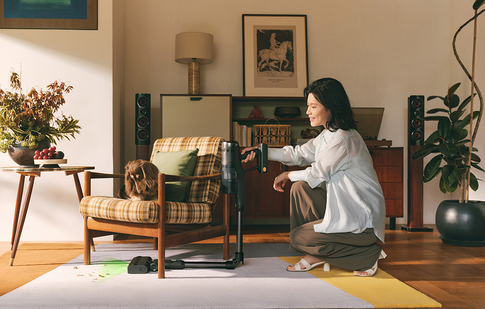 Kobieta używa odkurzacza. Pies siedzi na kraciastym fotelu. Po lewej stronie stół z kwiatami.