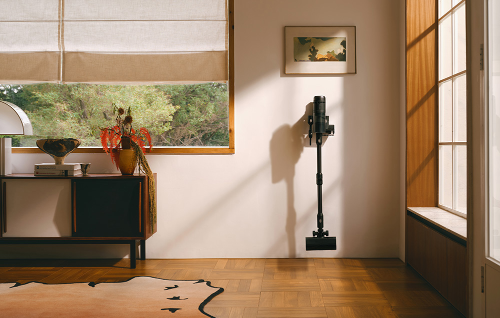 Czarny odkurzacz wisi na ścianie. W pokoju jest okno z żaluzjami i dywan. Widoczna jest szafka z dekoracjami.