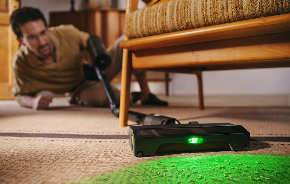 Mężczyzna odkurza pod krzesłem. Czarna głowica odkurzacza z zielonym światłem na dywanie.