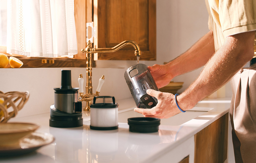 Osoba płucze element odkurzacza pod kranem. Otoczenie kuchni z innymi elementami na blacie i oknem.