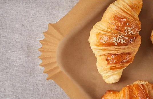 Zbliżenie rogalika z ziarnami sezamu na brązowej papierowej tacy. Tło to szara tkanina.