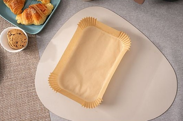 Pusta papierowa forma do pieczenia leży na beżowej podkładce. Rogaliki i ciastka obok na niebieskich i białych talerzach.