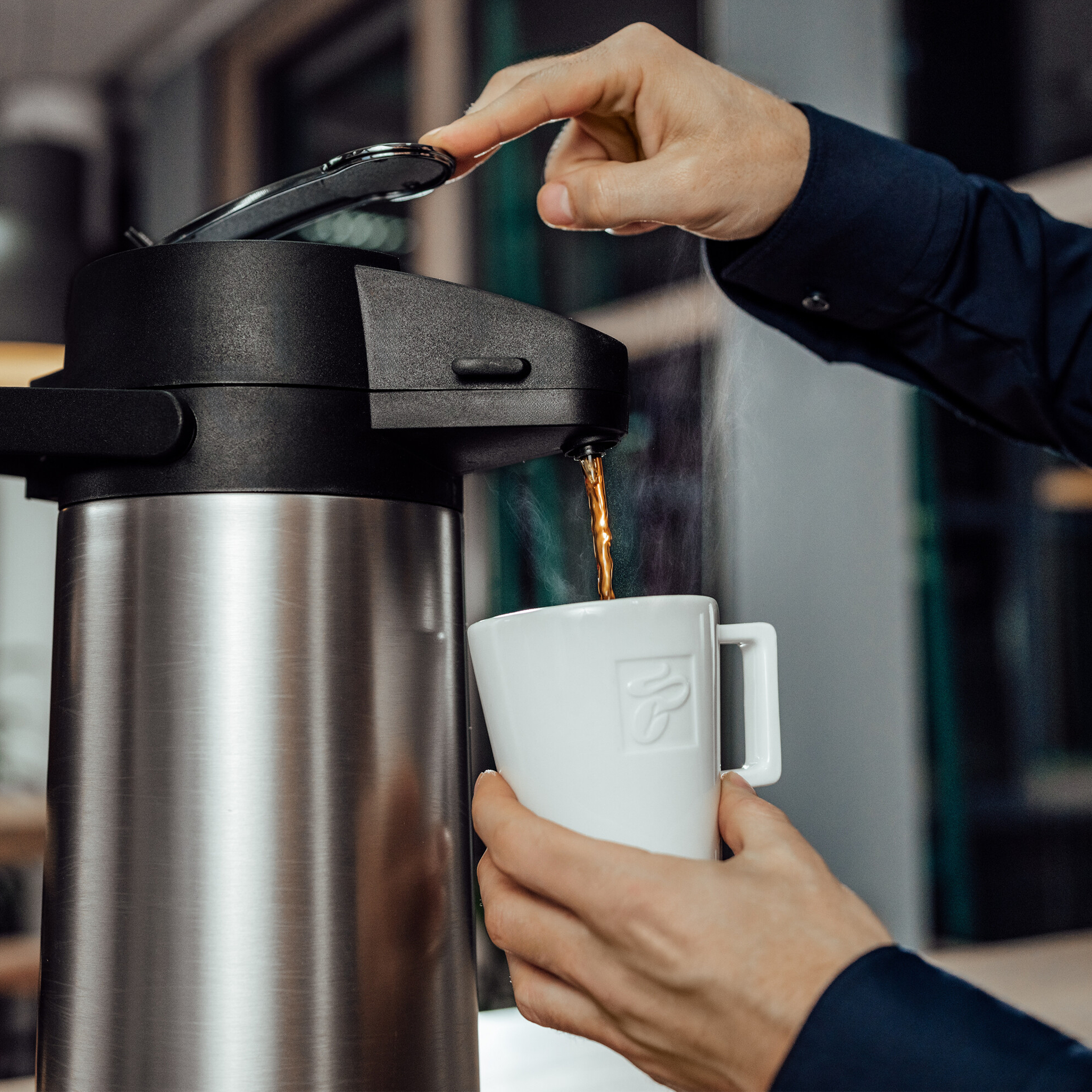 Eine Person gießt Kaffee aus einer Thermoskanne in eine weiße Tasse. Die Szene ist drinnen, mit verschwommenem Hintergrund.