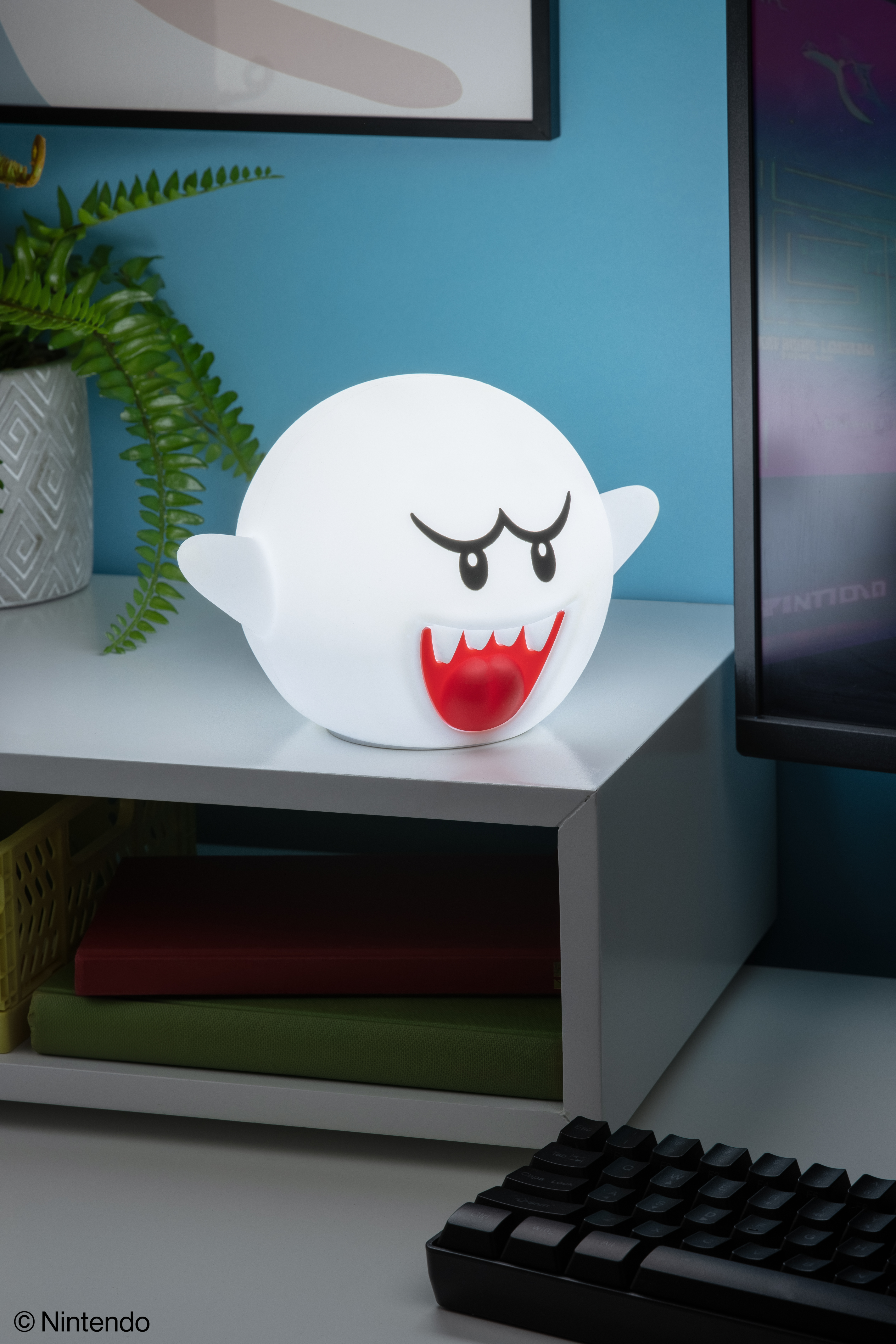 Eine weiße Boo-Lampe mit rotem Mund steht auf einem Regal. Grüne Pflanze, Bücher und eine Tastatur sind ebenfalls vorhanden. Blaue Wand im Hintergrund.