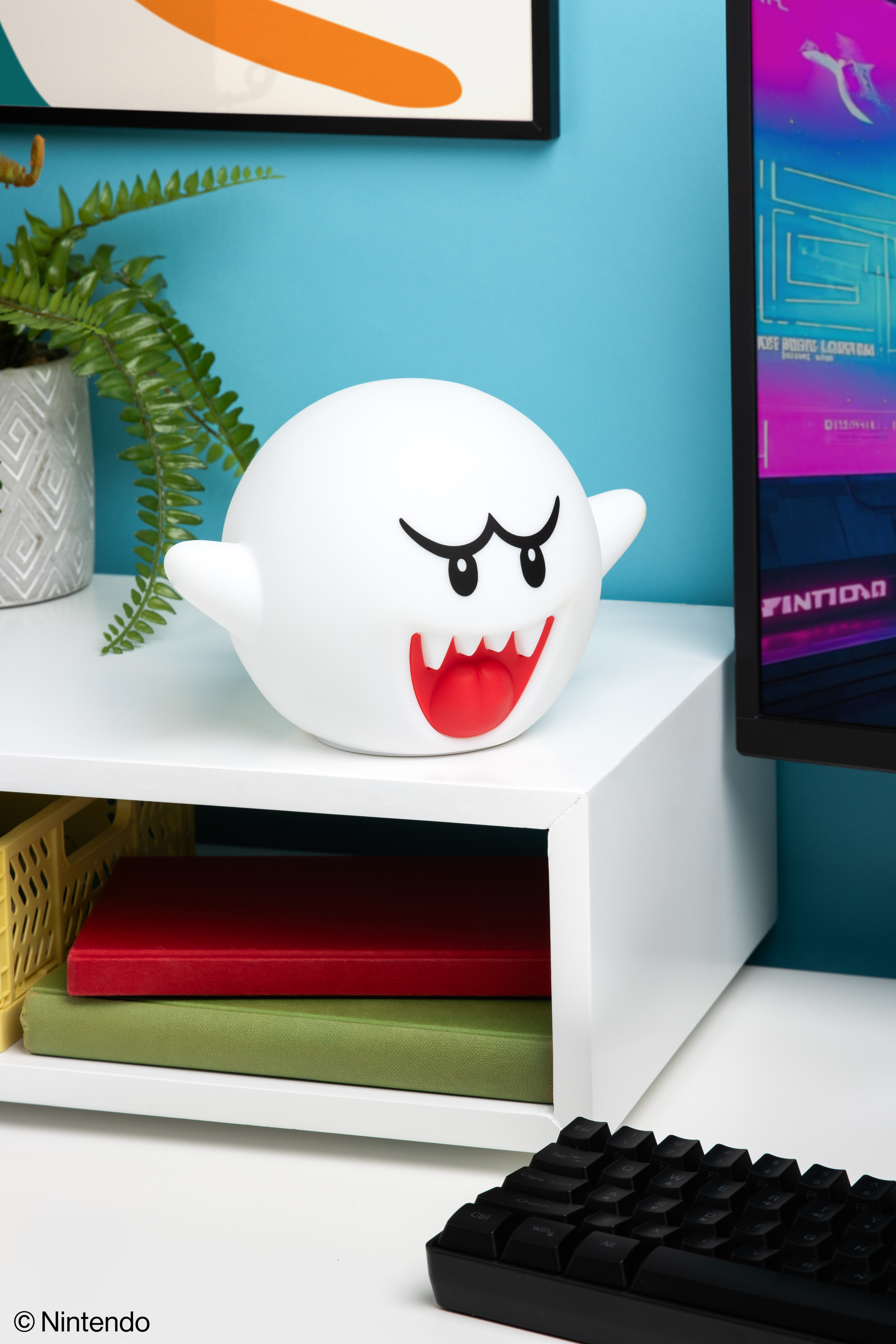 Eine weiße Boo-Lampe mit schwarzen Augen und Mund steht auf einem weißen Regal. Eine Tastatur ist darunter. Bücher und ein Farn sind vorhanden.