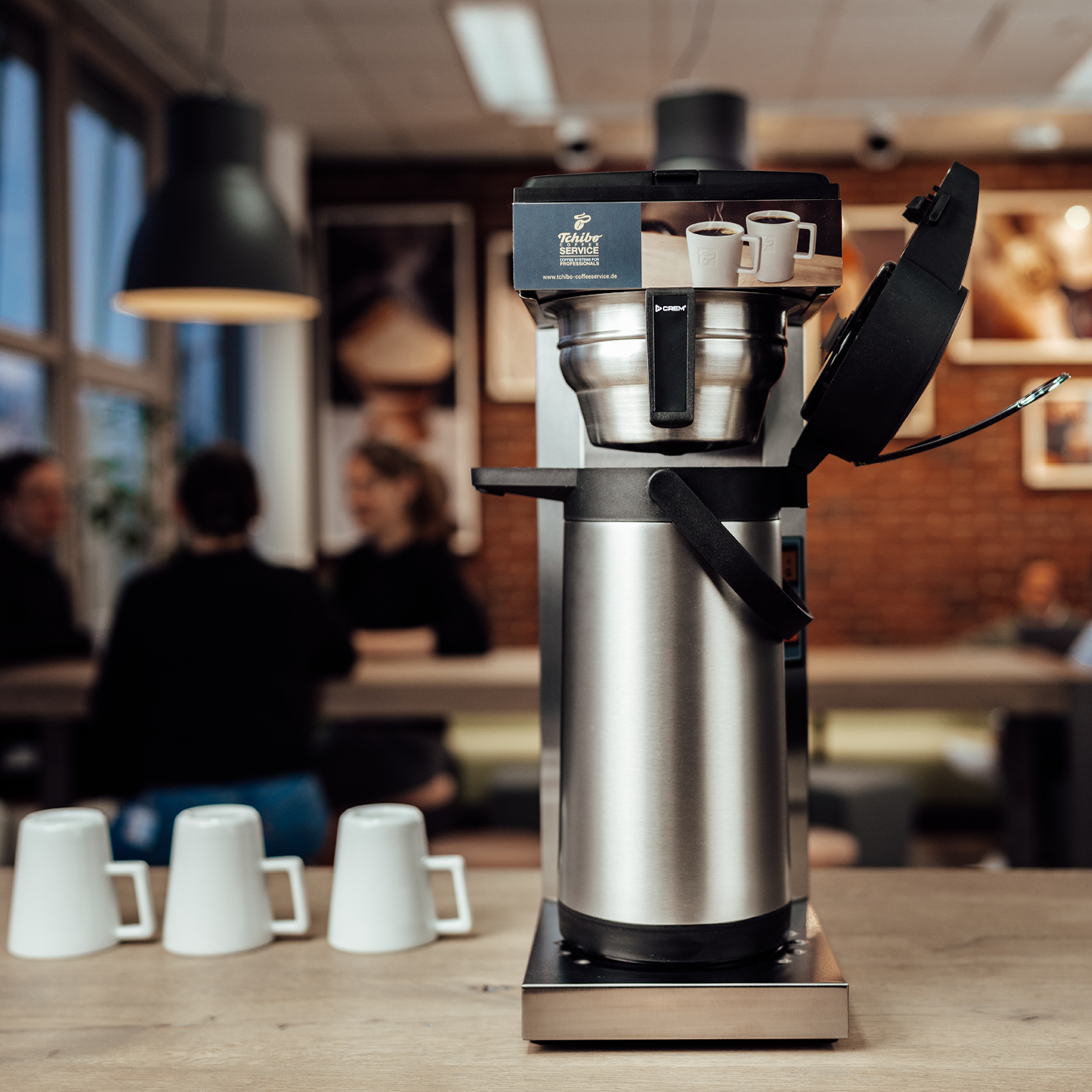 Kaffeemaschine mit silberner Karaffe und schwarzen Akzenten. Drei weiße Tassen stehen davor.