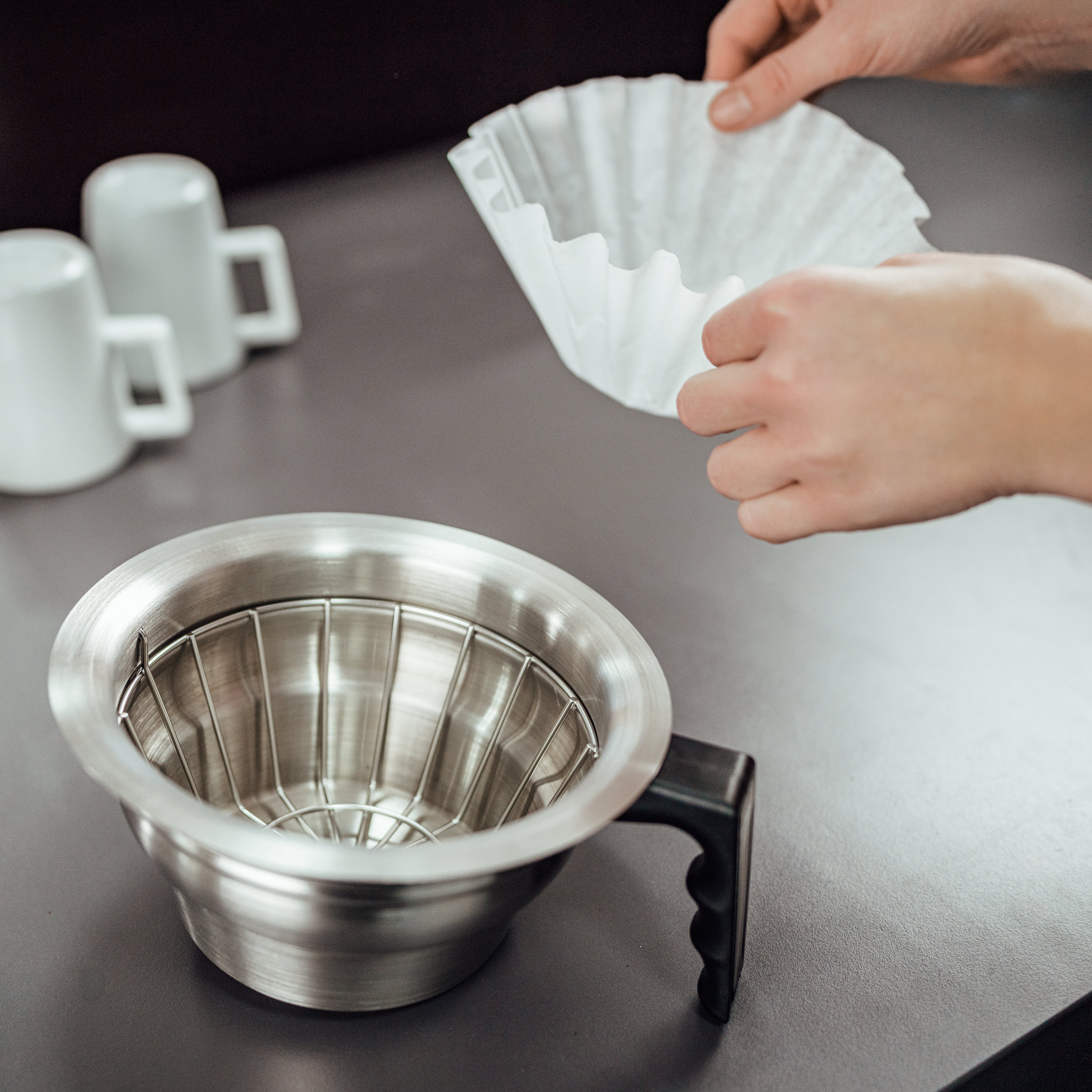 Hände halten einen weißen Papierfilter über einem Edelstahl-Kaffeefilterhalter. Links sind zwei weiße Tassen.