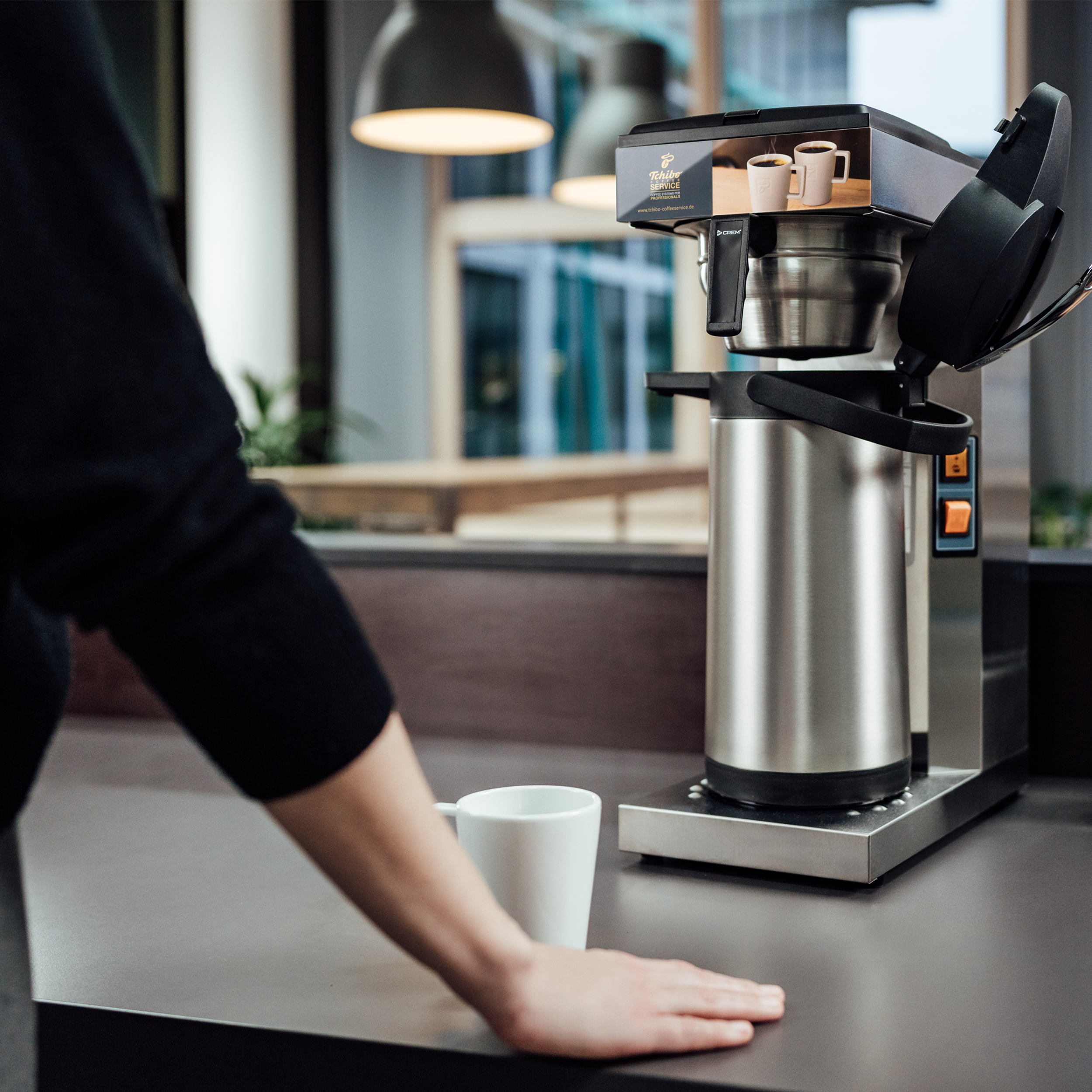 Die Hand einer Person in der Nähe einer Kaffeemaschine. Ein weißer Becher steht auf der Theke. Zwei Kaffeetassen auf der Maschine.