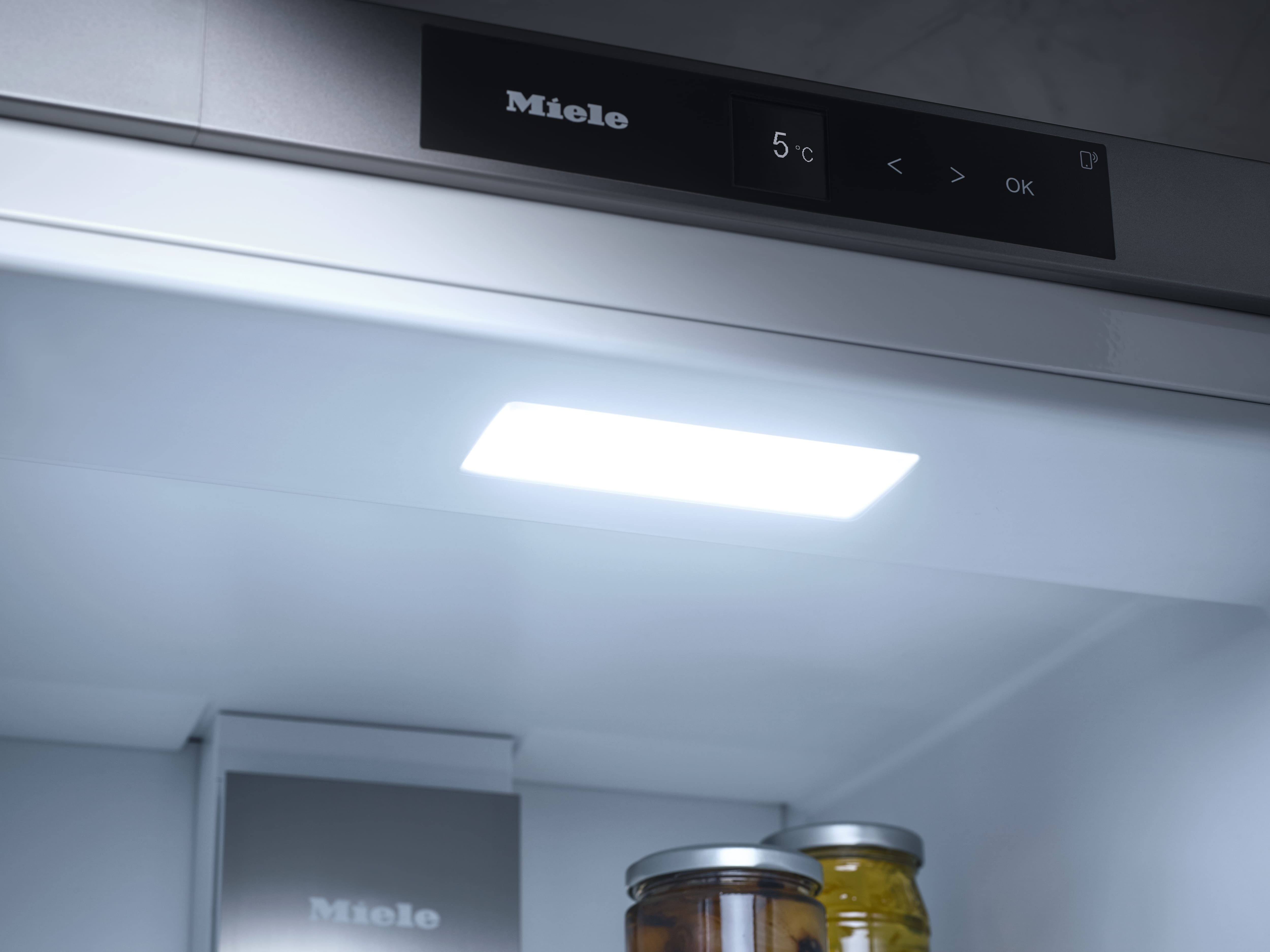 Im Kühlschrank. Ein helles Licht scheint von einem rechteckigen Panel. Gläser mit Lebensmitteln sind unten sichtbar. Ein Panel zeigt 'Miele' und Temperatur.