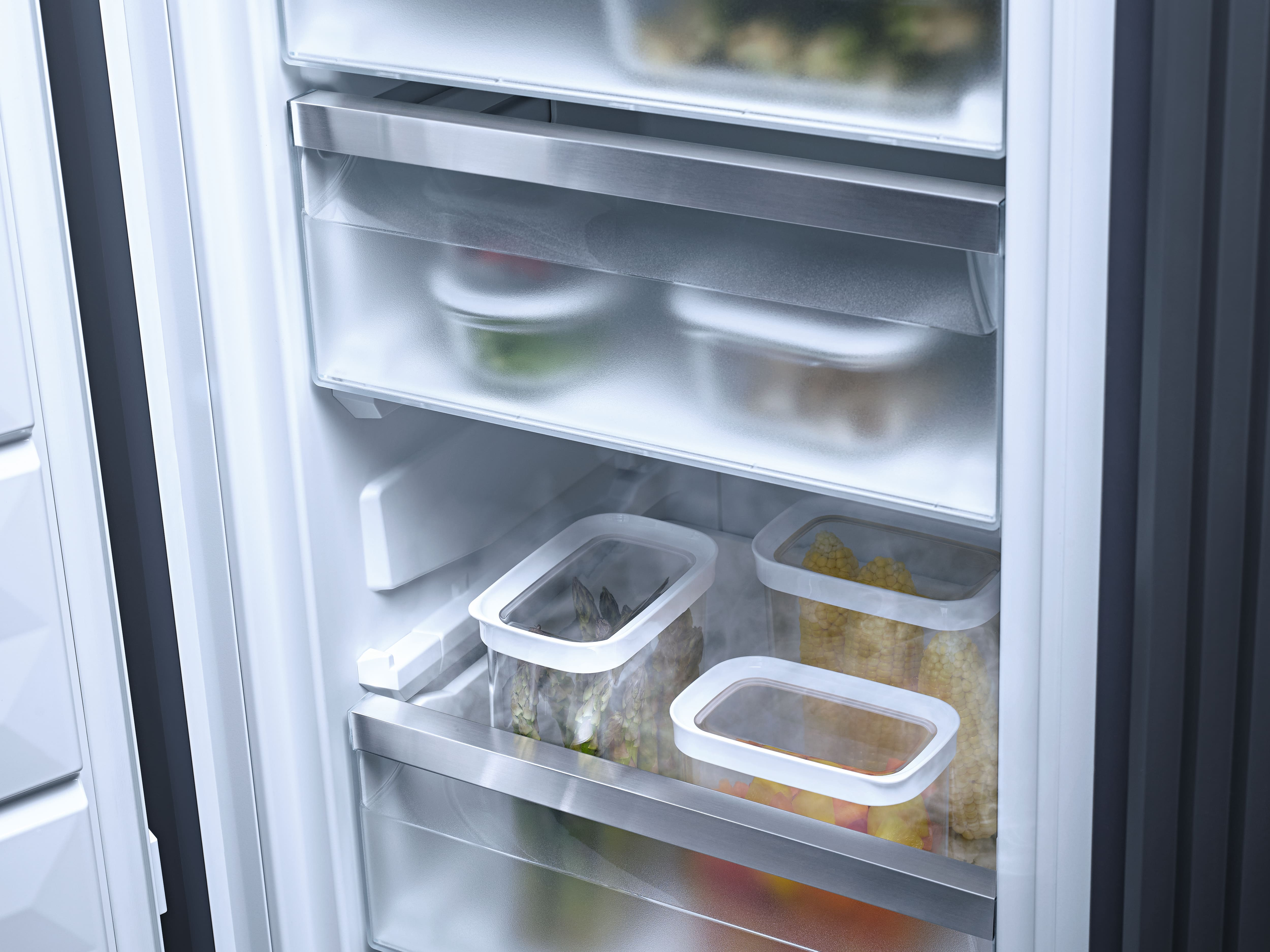 In einem Kühlschrank befinden sich transparente Behälter mit Gemüse und Mais. Die Regale sind aus Stahl, die Tür ist weiß und grau.