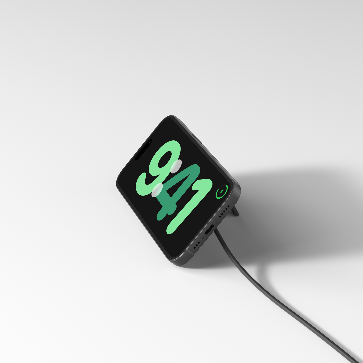 Un smartphone noir affiche 9h41 en vert. Il se recharge avec un câble noir sur une surface blanche, avec une ombre.