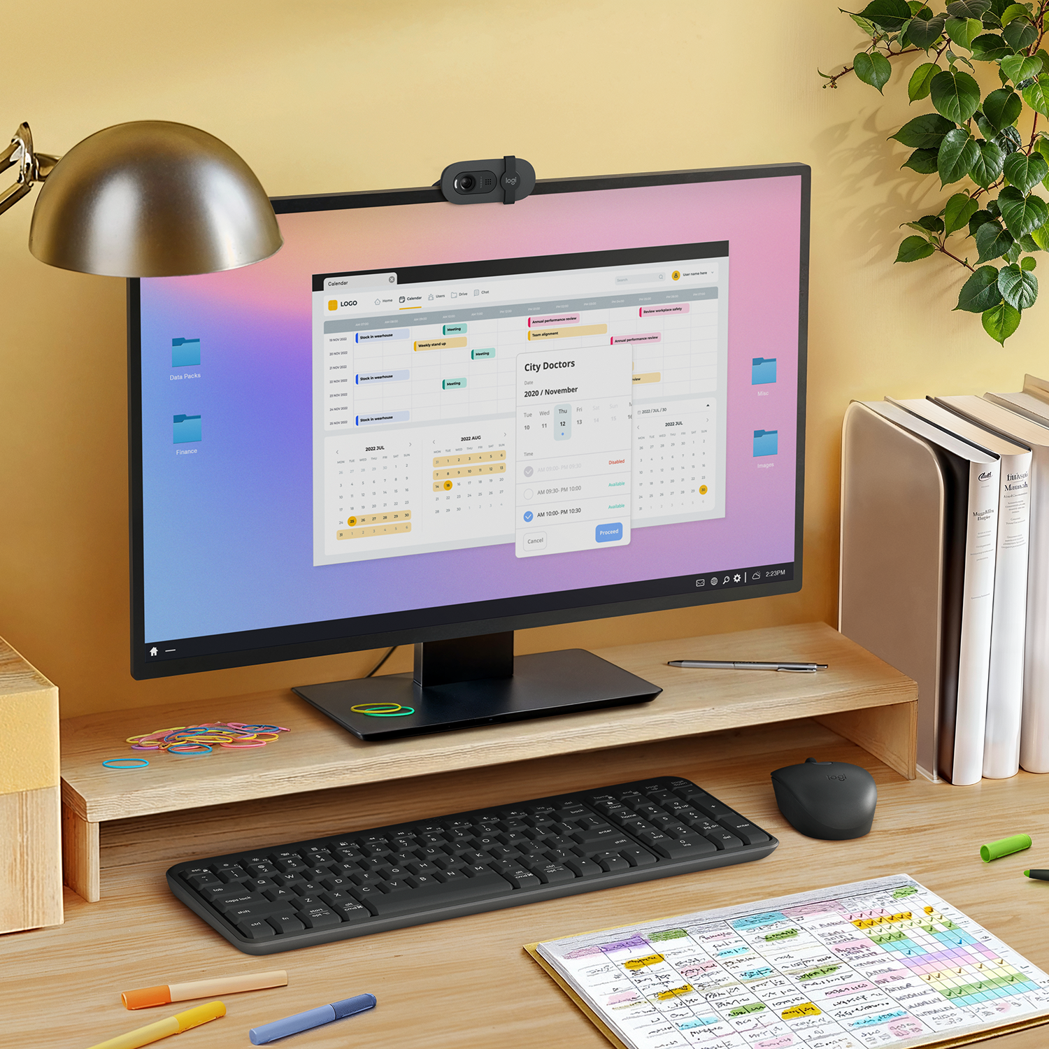 Una configuración de escritorio incluye un monitor, teclado, ratón y un calendario. Hay una webcam en la parte superior del monitor, con artículos de papelería y libros a la derecha.