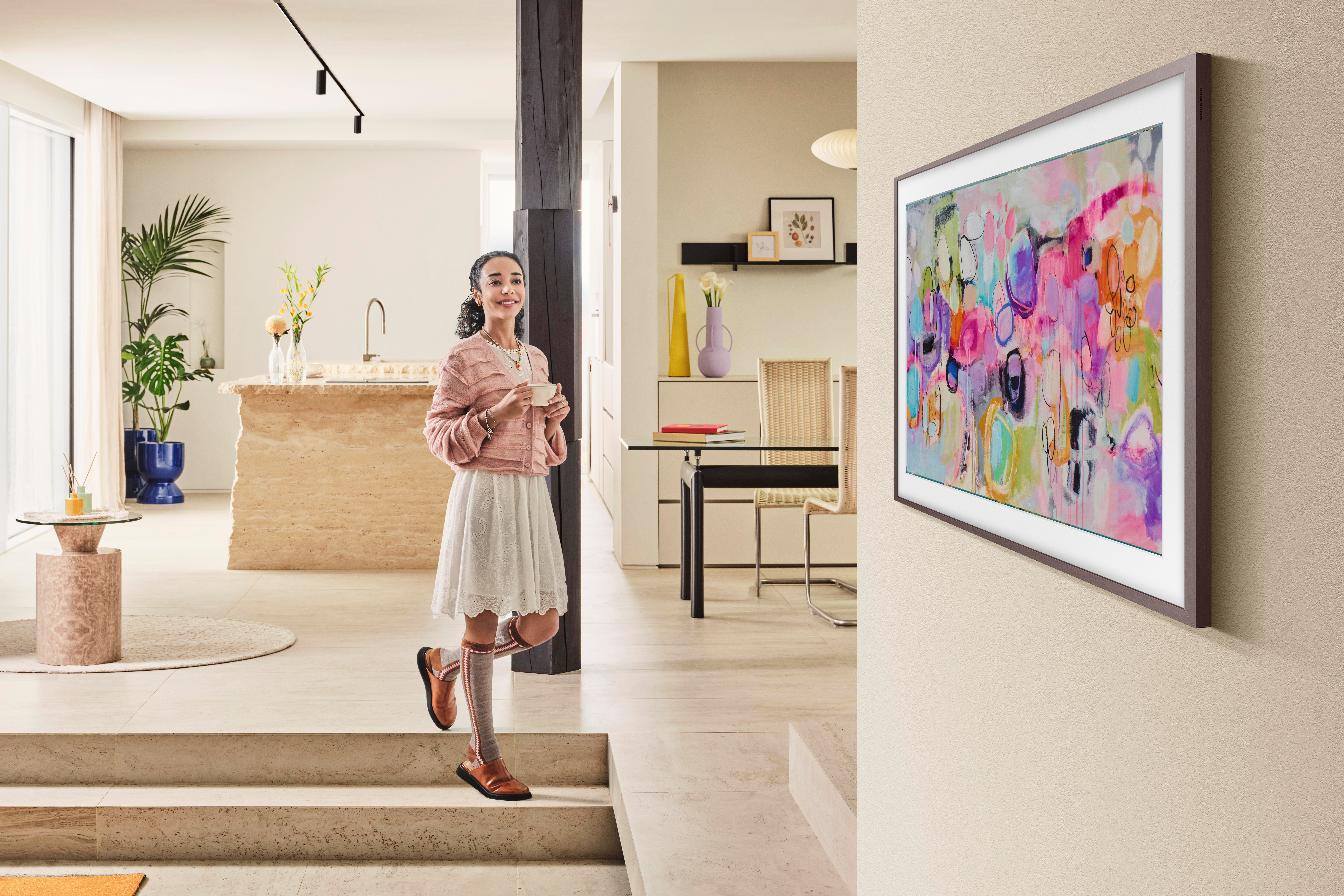 Frau mit rosa Strickjacke und weißem Rock hält eine Tasse und lächelt. Abstrakte Kunst an der Wand. Helles, modernes Interieur.