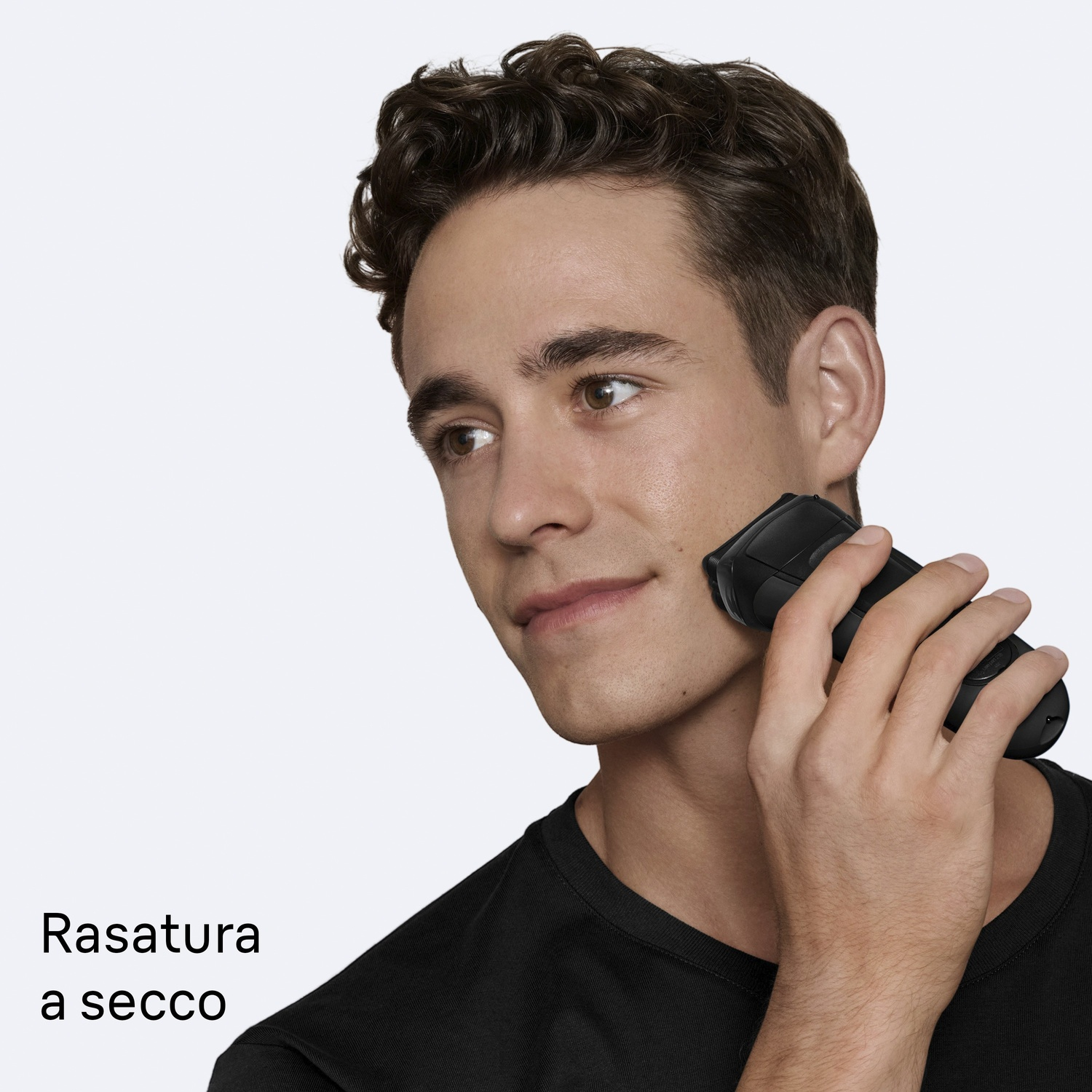 Un uomo si rade con un rasoio elettrico. Indossa una maglietta nera su sfondo bianco. Testo italiano: Rasatura a secco.