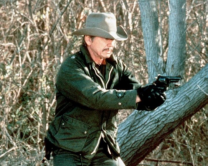 Ein Mann mit Hut und grüner Jacke zielt mit einer Waffe. Er steht in der Nähe von Bäumen. Der Hintergrund hat verschwommene Äste und Blätter.
