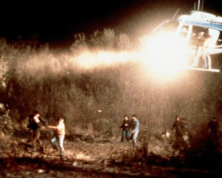 Ein Hubschrauber beleuchtet Menschen in einem Feld bei Nacht. Mehrere Figuren stehen im Vordergrund.