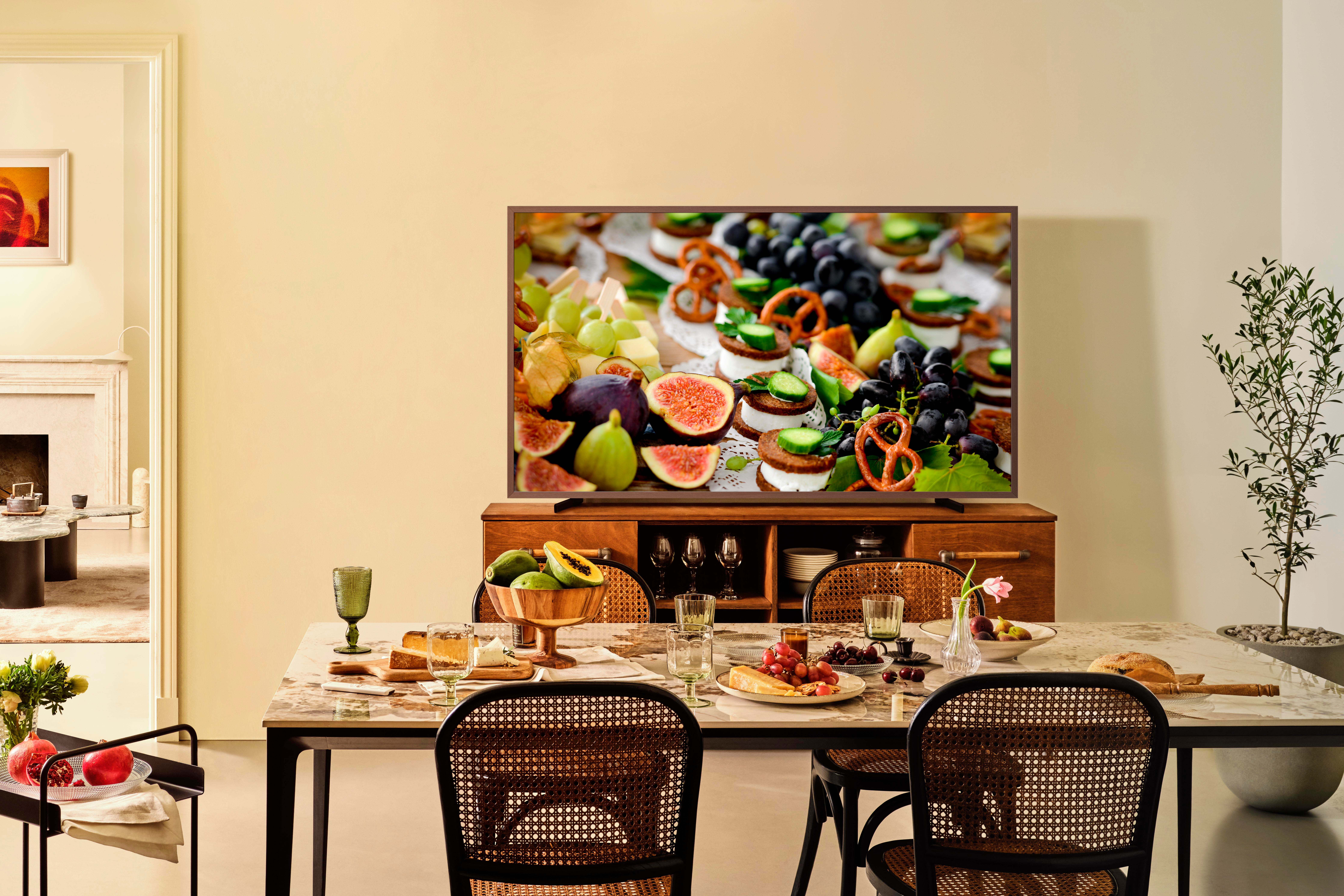 Ein Esszimmer mit gedecktem Tisch. Der Fernseher zeigt Essen, darunter Feigen und Trauben. Stühle stehen am Tisch.