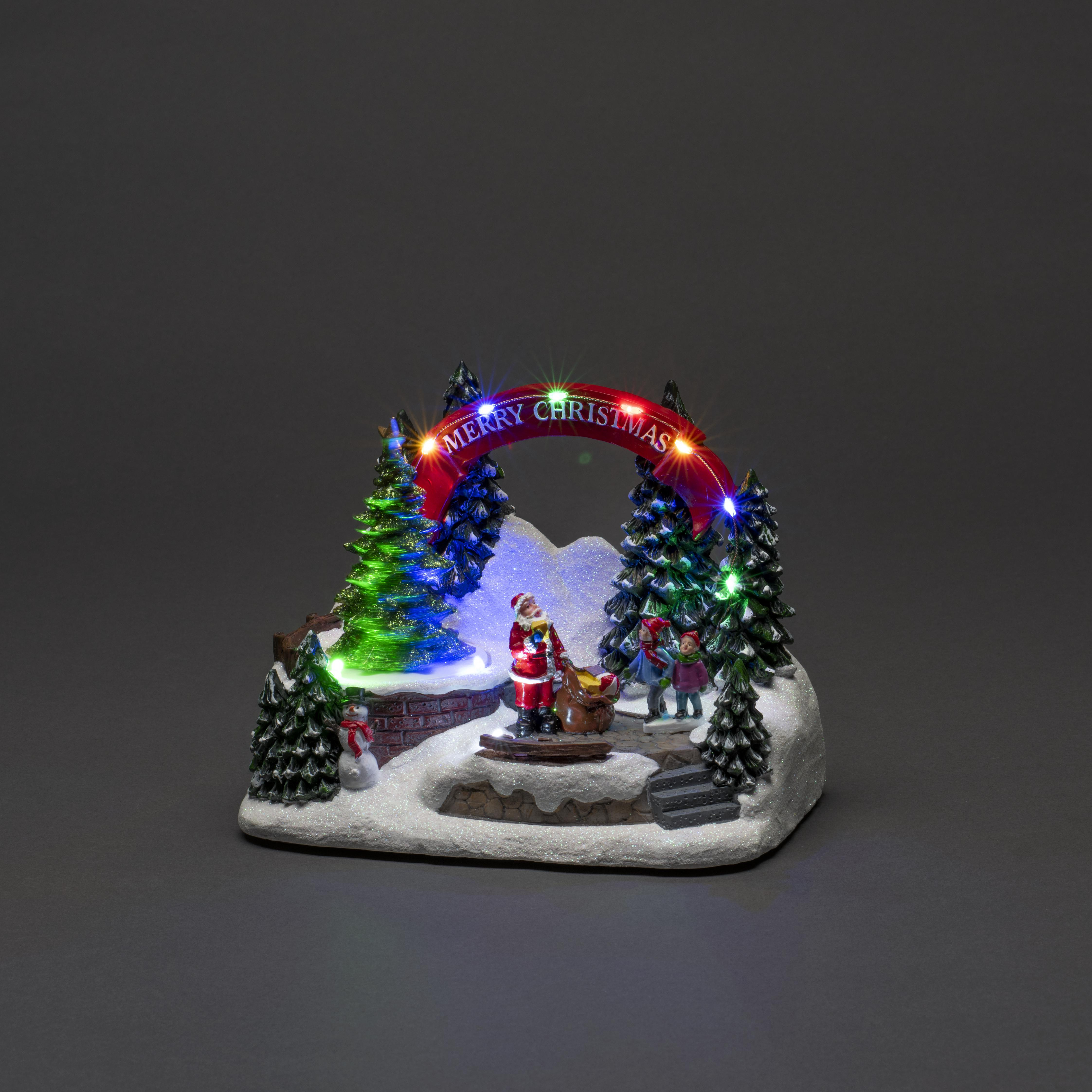 Eine Weihnachtsszene mit Weihnachtsmann, Kindern und einem beleuchteten 'Merry Christmas'-Bogen. Schnee, Bäume und ein Schneemann.