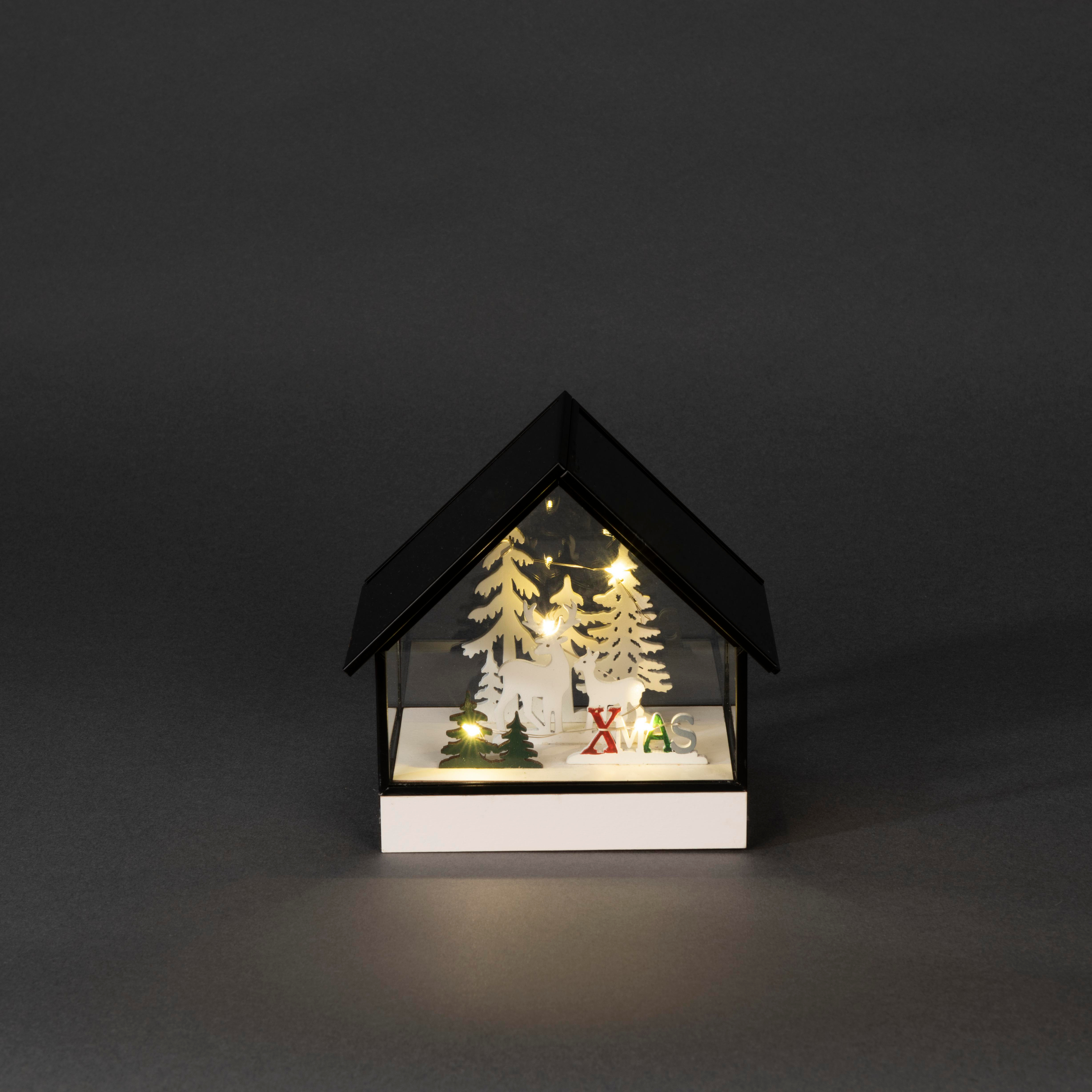 Ein hausförmiges Weihnachtsdekor. Im Inneren sind Bäume, Hirsche und XMAS-Buchstaben beleuchtet. Schwarzes Dach, weiße Basis, grauer Hintergrund.