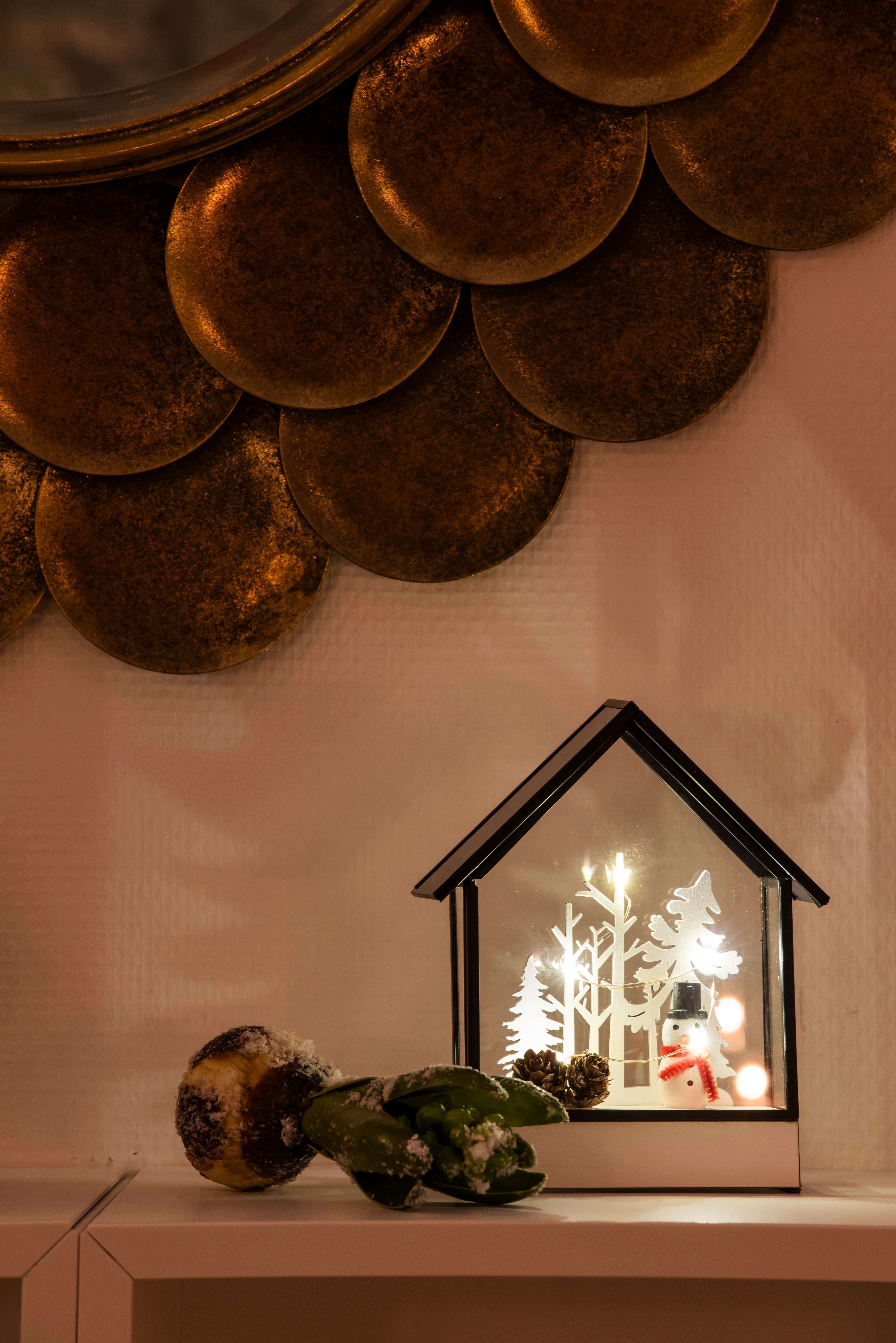Weihnachtsszene mit Deko-Haus, Schneemann und Blättern. Darüber ein goldenes Kreisdesign. Die Szene ist warm beleuchtet.