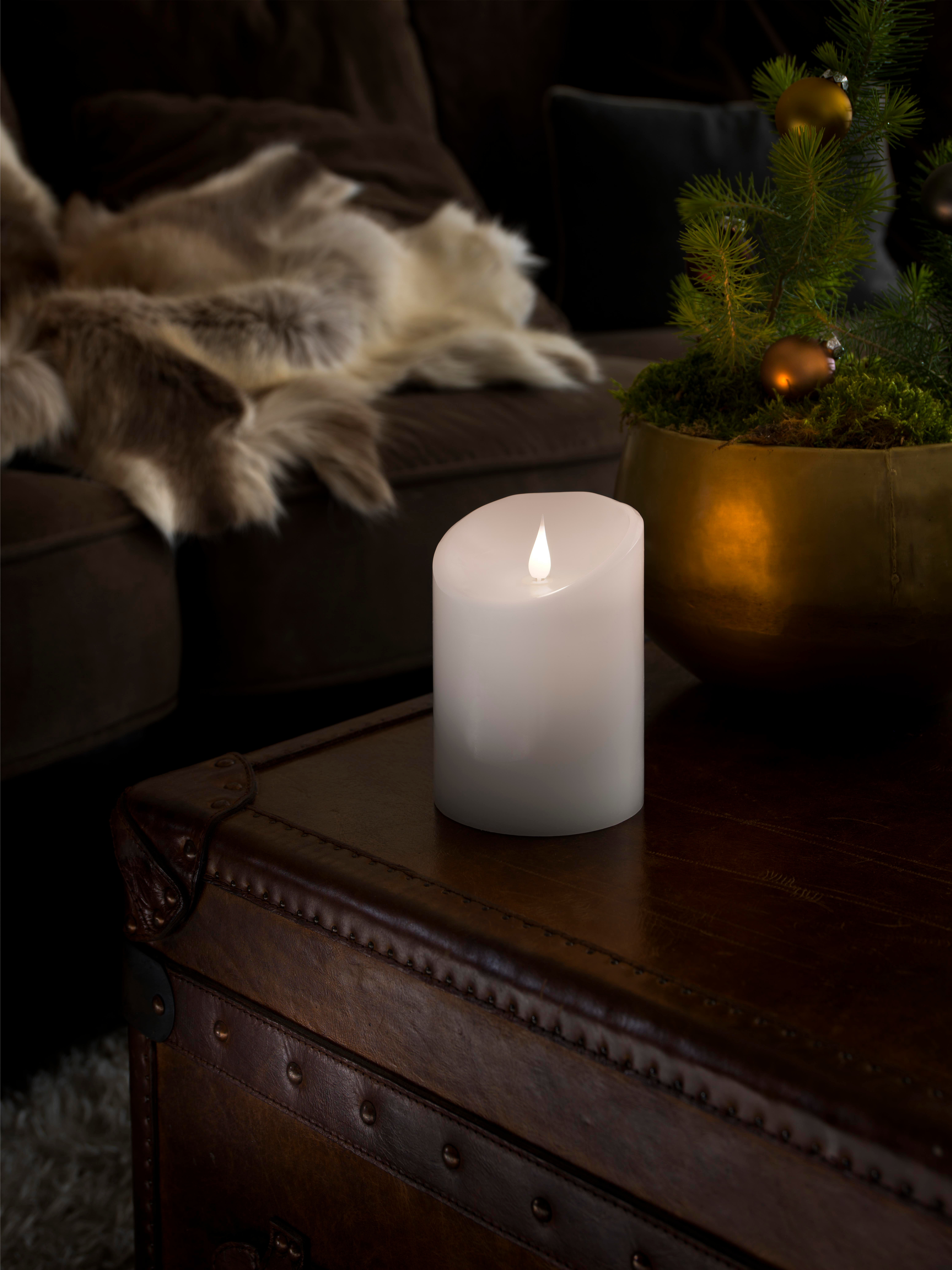 Eine weiße Kerze mit flackernder Flamme steht auf einem dunkelbraunen Koffer. Ein kleiner Weihnachtsbaum steht in einem goldenen Topf.