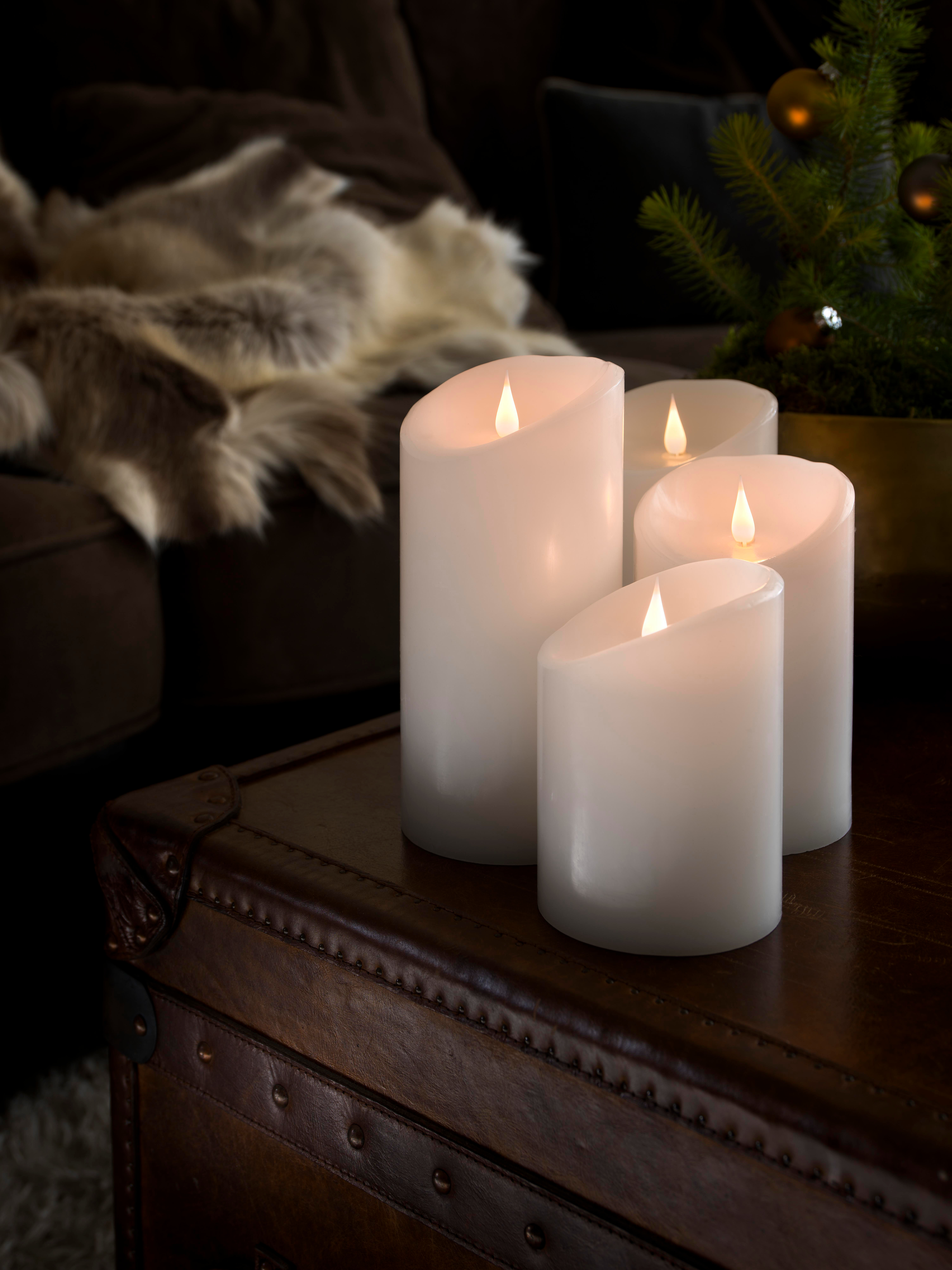 Vier weiße Kerzen mit Flammen auf einem braunen Lederkoffer. Ein Weihnachtsbaum und eine braune Couch sind im Hintergrund.