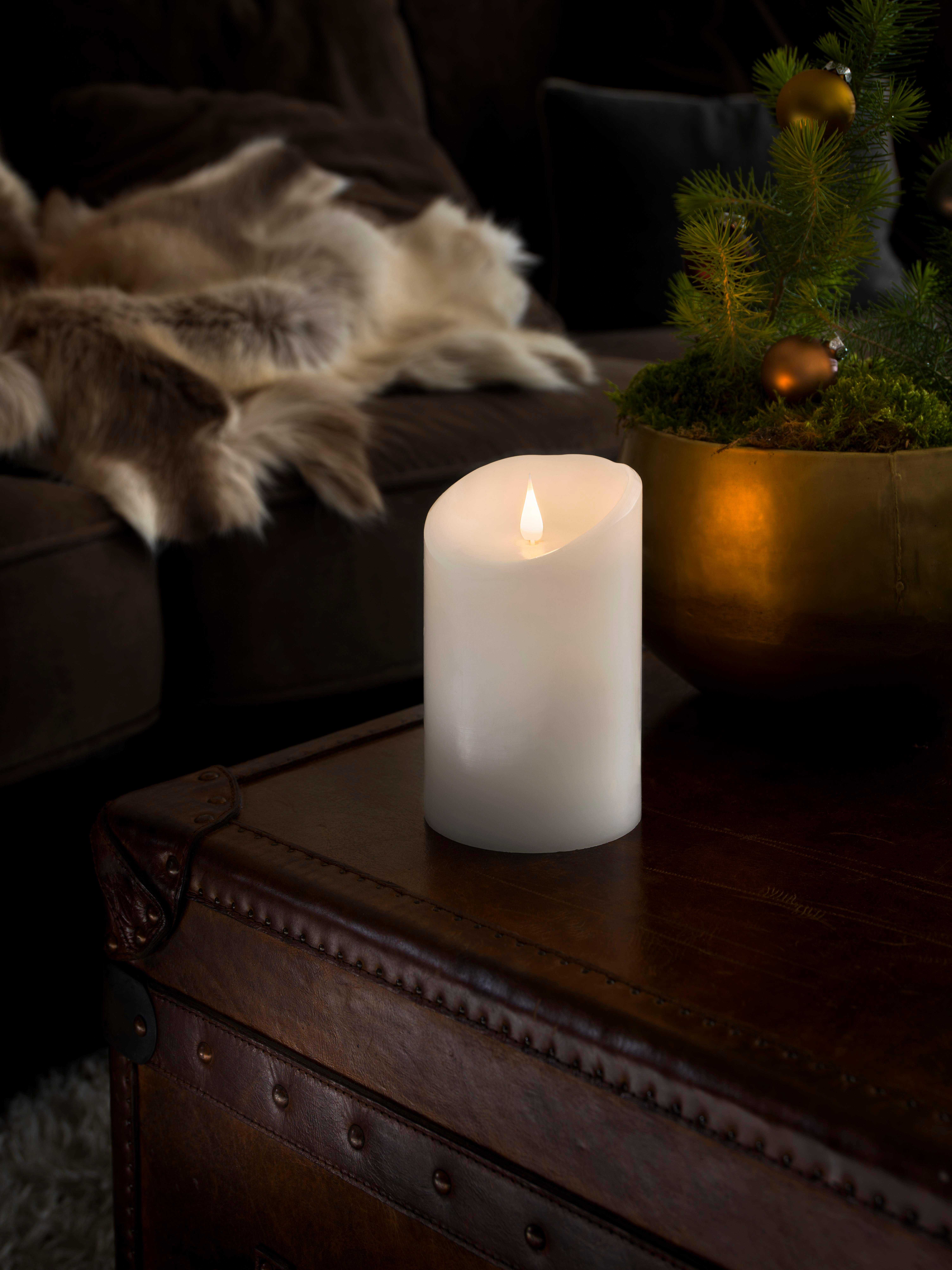 Eine weiße Kerze mit Flamme steht auf einer braunen Ledertruhe. Im Hintergrund ein kleiner Weihnachtsbaum.