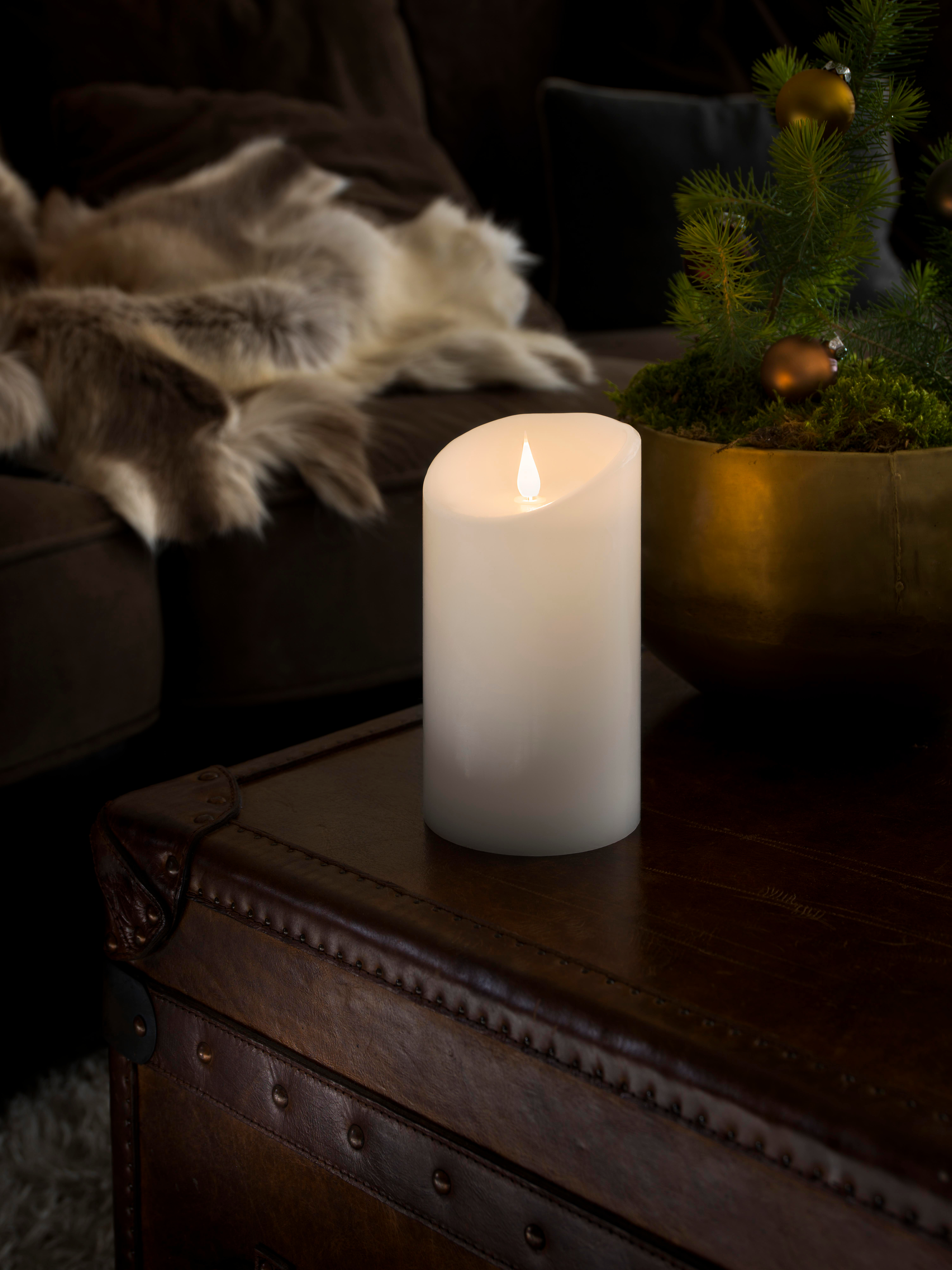 Eine brennende weiße Kerze auf einem braunen Koffer, ein Weihnachtsbaum in einer goldenen Schale und eine Pelzdecke im Hintergrund.