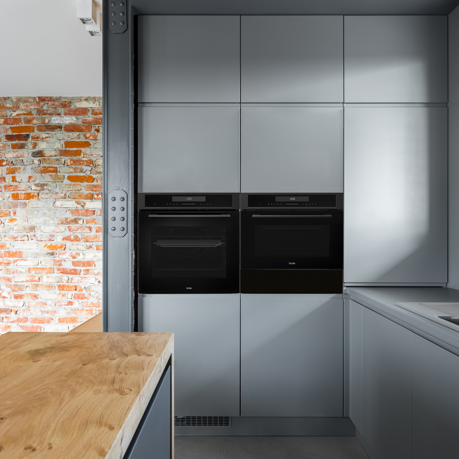 Modern kitchen with gray cabinets, two black ovens, and a wooden countertop. A brick wall and metal beam are visible.