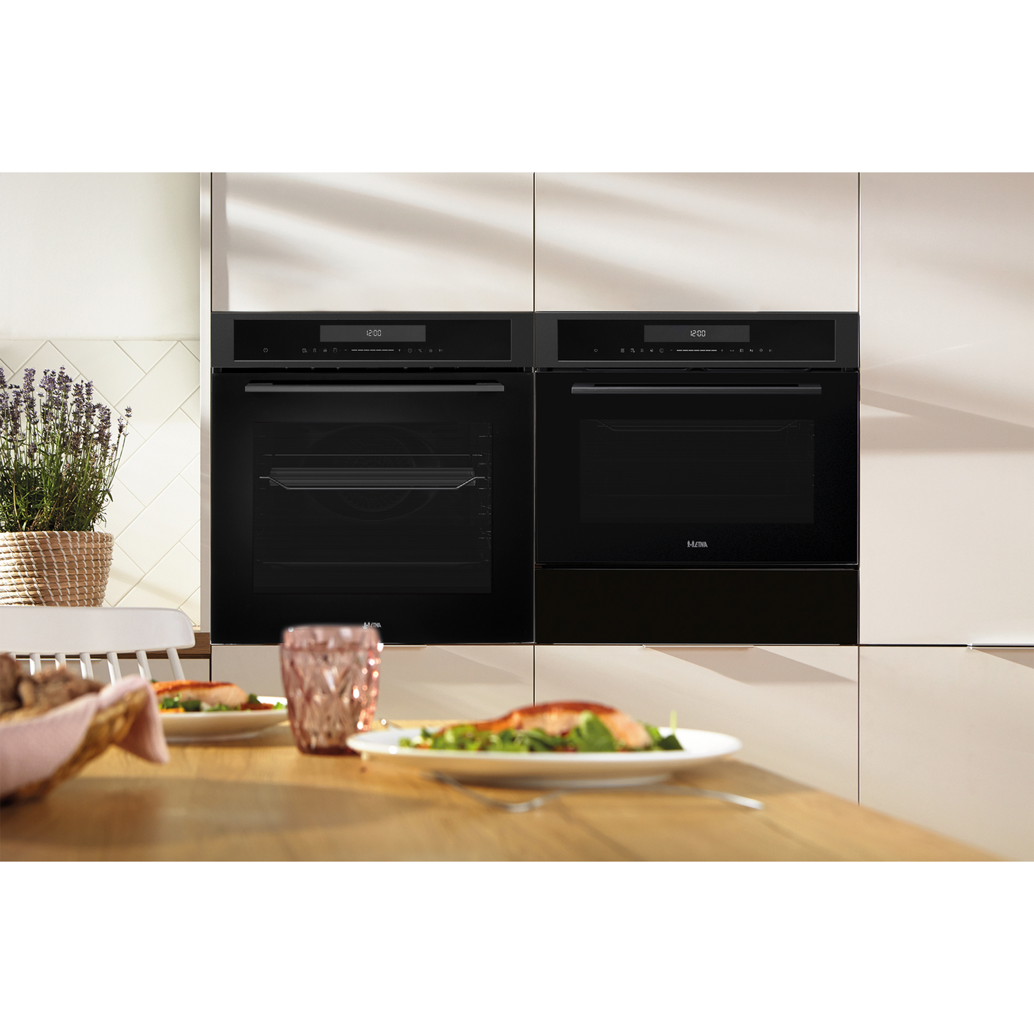 Two black built-in ovens with digital displays. A wooden table with plates of food, and a potted plant are in the foreground.