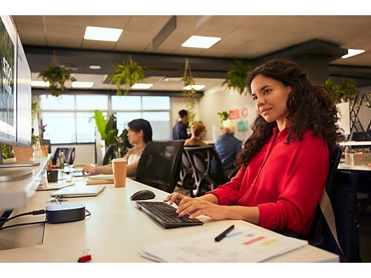 A woman in a red shirt types on a keyboard at her desk in an office. Other people work in the background.