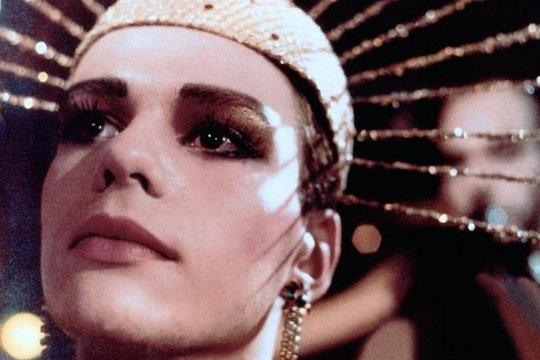 Eine Person mit Make-up, die ein geschmücktes Kopfstück und Ohrringe trägt, blickt nach oben. Weichzeichner im Hintergrund mit einer anderen Person.