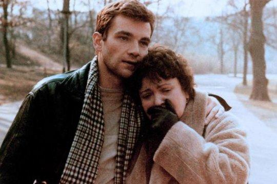 Ein Mann tröstet eine weinende Frau. Er trägt Jacke und Schal. Sie trägt einen Mantel und Handschuhe. Bäume sind im Hintergrund.