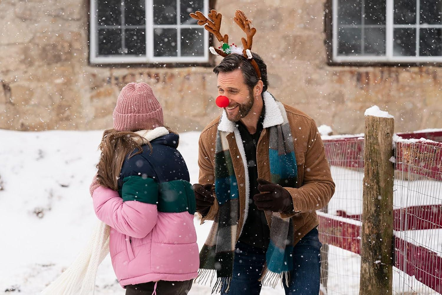 Ein Mann mit Rentiergeweih und roter Nase lächelt ein Kind in einem rosa Mantel an, schneebedeckter Hintergrund.