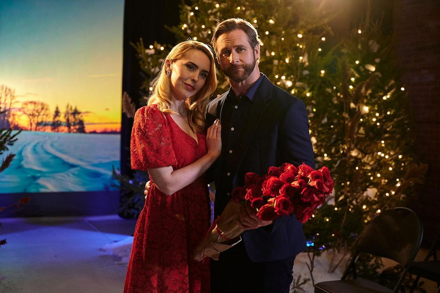 Ein Paar steht eng beieinander, die Frau in Rot, der Mann im Anzug mit Rosen. Im Hintergrund eine Winterlandschaft und ein Weihnachtsbaum.