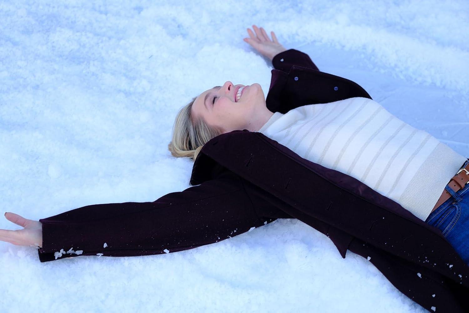 Eine lächelnde Frau liegt im Schnee. Sie trägt einen dunklen Mantel, einen weißen Pullover und Jeans. Arme ausgestreckt. Der Hintergrund ist Schnee.