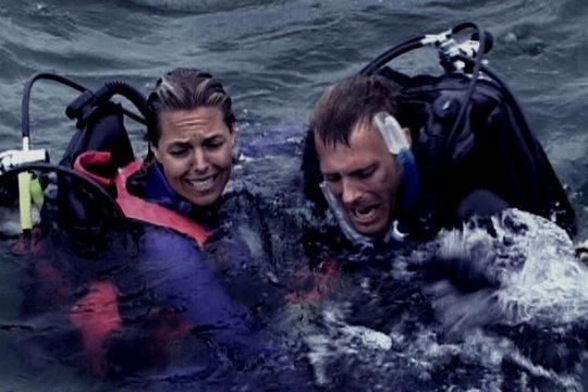 Zwei Taucher im Meer, die betrübt aussehen. Sie tragen Tauchausrüstung und sind von Wasser umgeben. Einer hat eine rote Schwimmweste.
