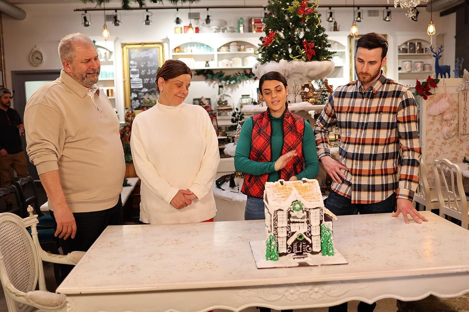 Vier Personen stehen um einen Tisch mit einem Lebkuchenhaus. Ein Weihnachtsbaum und Dekorationen befinden sich im Hintergrund.
