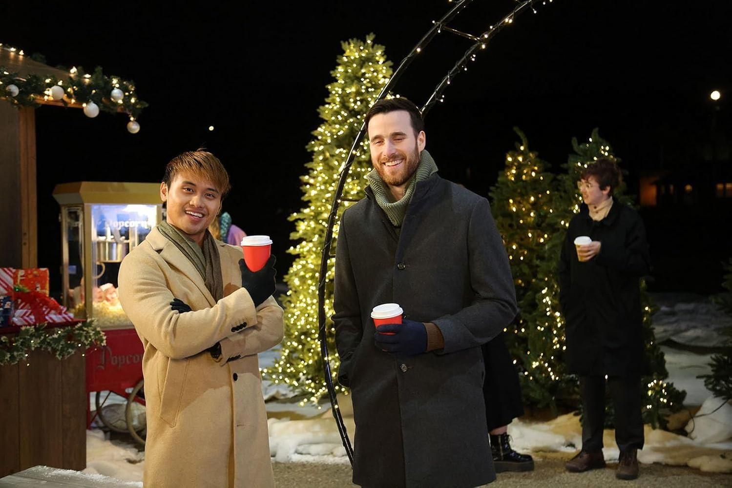 Drei Personen im Freien bei Nacht, die Kaffeetassen halten. Weihnachtsbäume und Lichter im Hintergrund. Popcorn-Stand links.