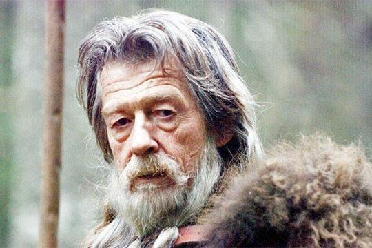 Ein älterer Mann mit langem, weißem Bart und Haaren, mit einem Pelzkragen, der in die Kamera schaut. Ein Holzstab ist an der Seite zu sehen.