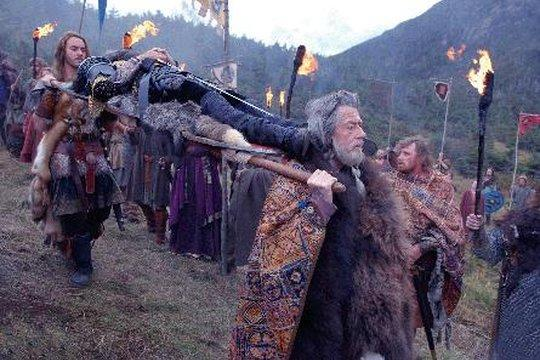 Menschen tragen eine Person auf einer Bahre. Sie tragen mittelalterliche Kleidung, halten Fackeln, Flaggen im Hintergrund.