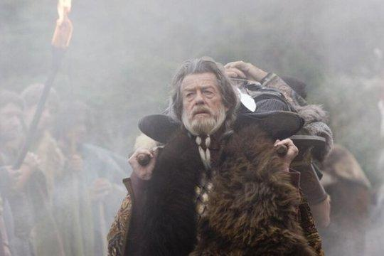 Ein alter Mann in Pelz trägt einen Körper auf einer Bahre. Viele Menschen mit Fackeln sind im Hintergrund, eingehüllt in Nebel.