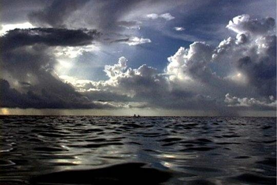 Dramatische Meereslandschaft mit dunklem Wasser und bewölktem Himmel. Sonnenlicht scheint durch die Wolken. Zwei kleine Boote am Horizont.