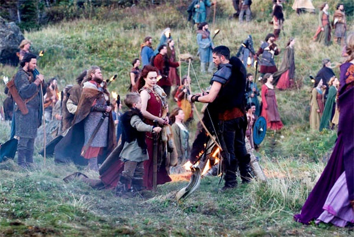 Eine Gruppe von Menschen in mittelalterlicher Kleidung steht im Freien. Davor brennt ein Feuer. Einige Leute halten Bögen und Pfeile. Viele Menschen sind im Hintergrund.