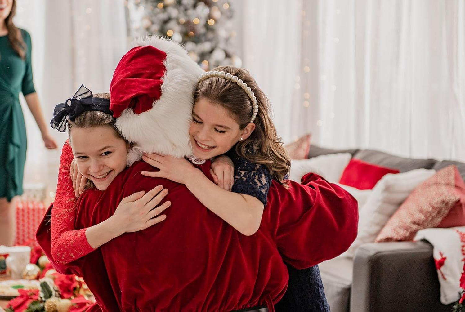 Zwei Mädchen umarmen den Weihnachtsmann. Eines trägt ein Perlenstirnband, das andere eine schwarze Schleife. Ein Weihnachtsbaum und Sofa im Hintergrund.