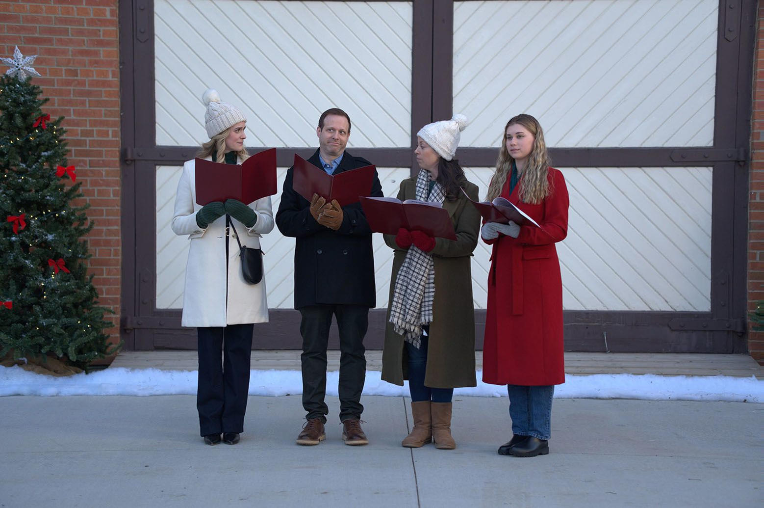 Vier Personen singen mit roten Büchern vor einem Gebäude und einem Weihnachtsbaum. Sie tragen Mäntel und Hüte. Schnee vorhanden.