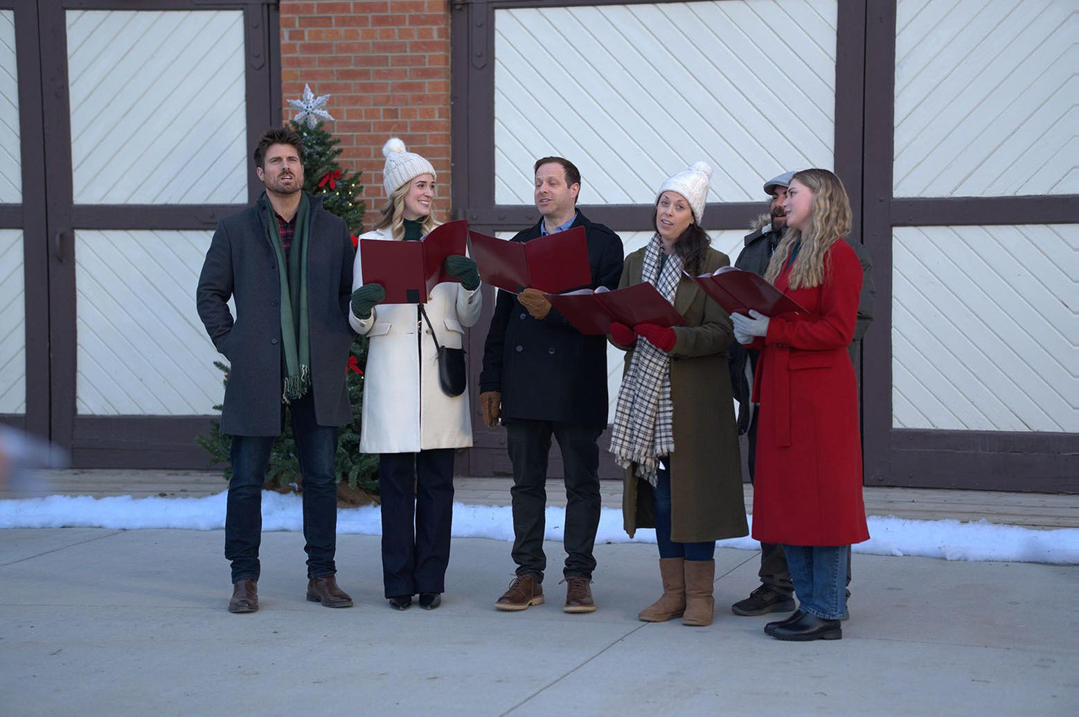 Eine Gruppe von Menschen singt im Freien Weihnachtslieder. Sie tragen Mäntel, Mützen und Schals. Im Hintergrund ist ein Weihnachtsbaum zu sehen. Ein Gebäude ist ebenfalls vorhanden.